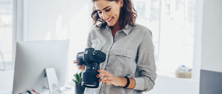 Female photographer checking pictures on camera and smiling. Caucasian woman in casuals standing in her office looking at the photos on her dslr camera.